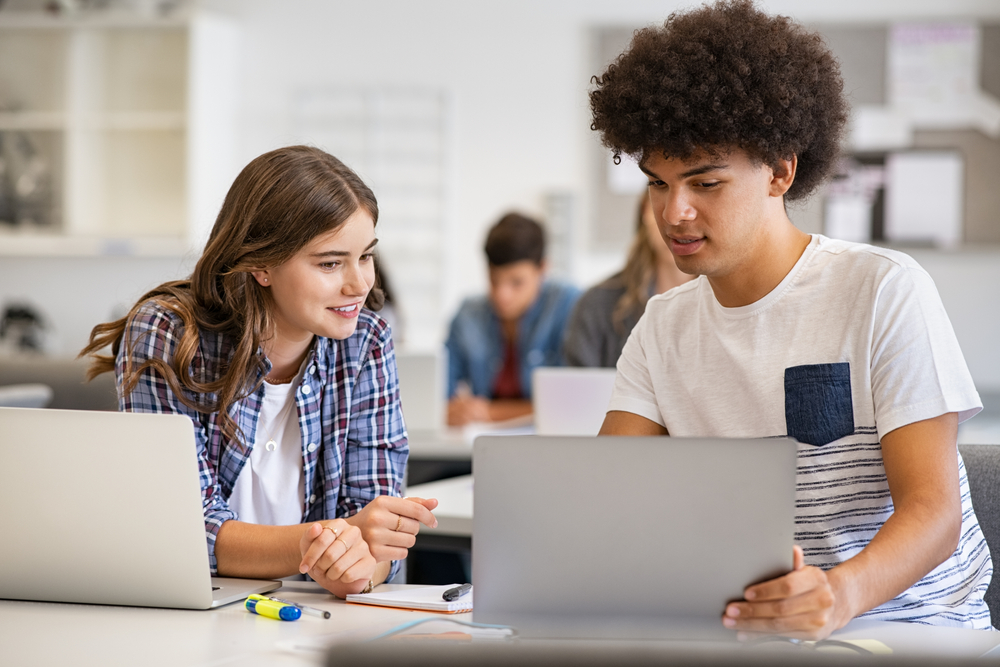 um aluno adolescente mostra a tela de um laptop para outra aluna, sentados em sala de aula.