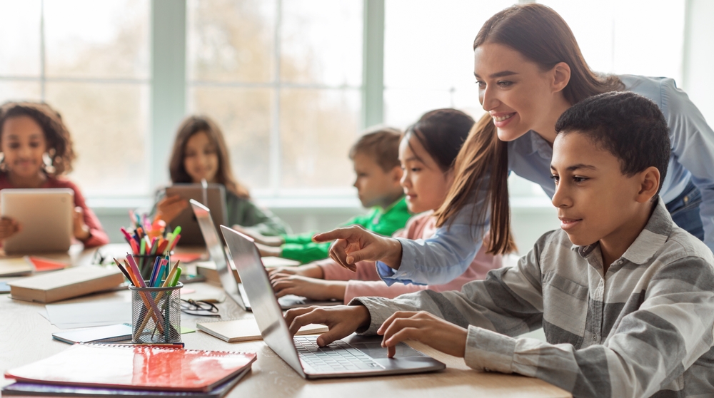 Em sala de aula bastante iluminada com janelas grandes ao fundo, alunos pequenos usam laptops com a ajuda da professora.