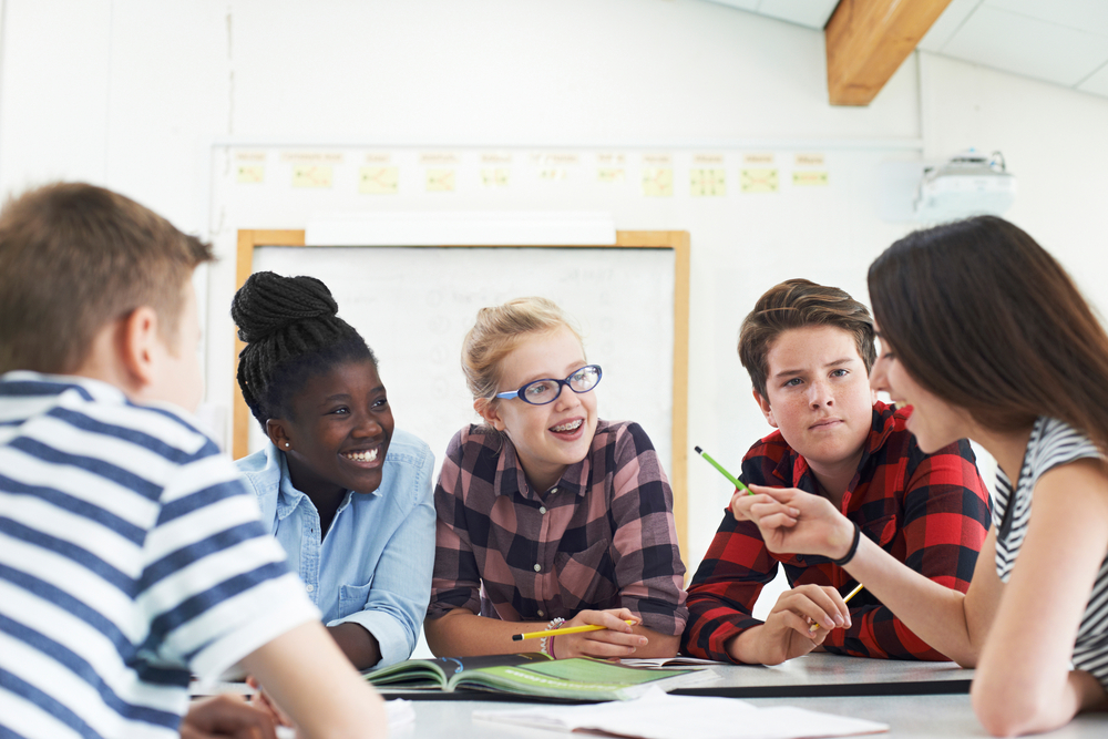 Grupo de cinco estudantes adolescentes reunidos ao redor de uma mesa, fazendo um trabalho em grupo.