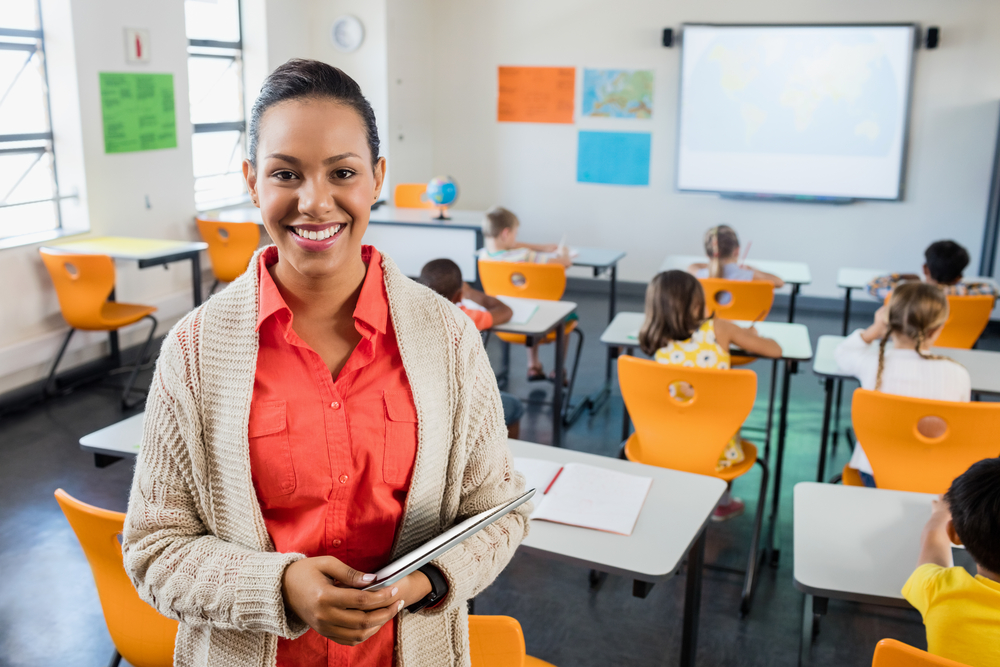 Professora negra de pele clara, cabelo preso, veste uma camisa salmão e cardigan bege claro. Ela sorri para a câmera, de pé, no meio da sala de aula, segurando um tablet nos braços. Ao fundo, crianças estão sentadas de costas nas carteiras.