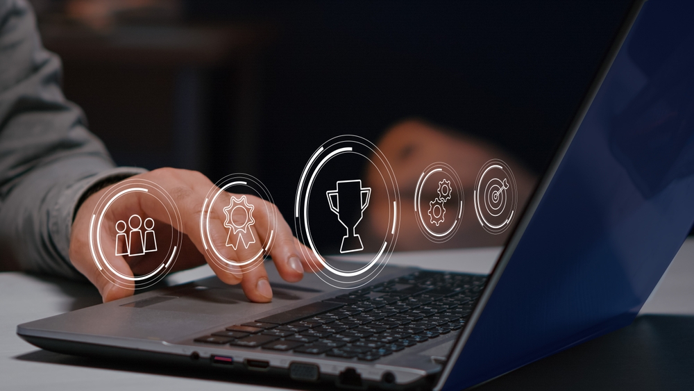 Mão de pessoa utilizando notebook com ícones de gamificação flutuando sobre a tela.