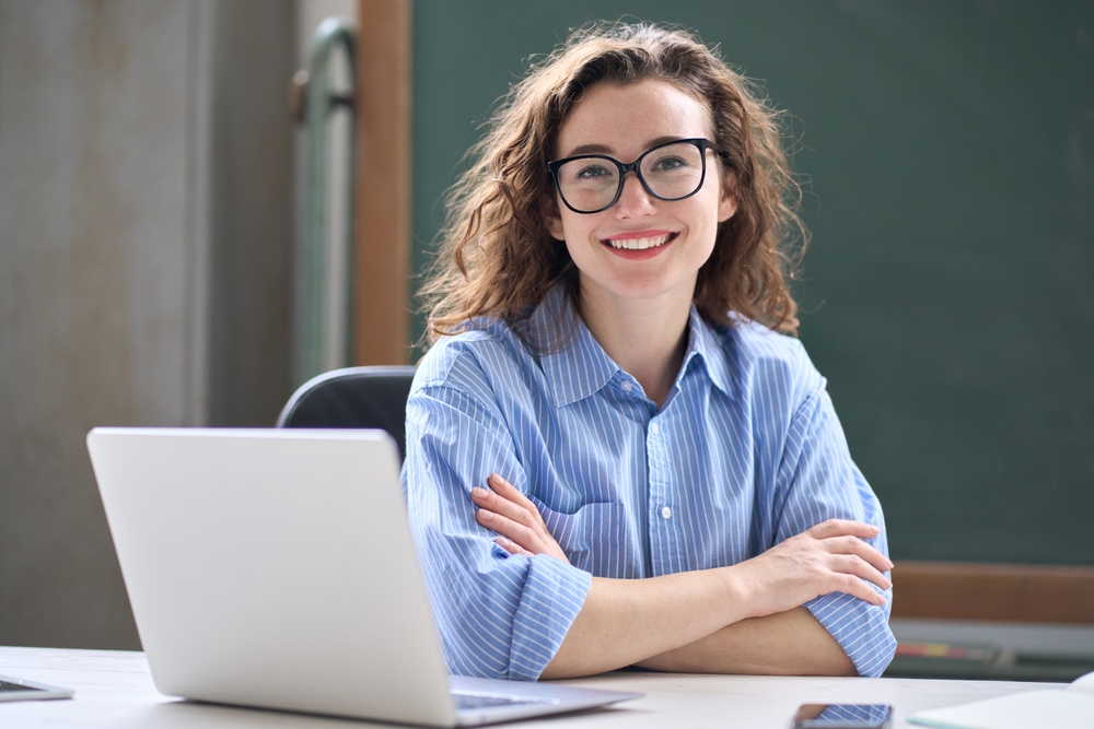Professora sentada em frente a um notebook, sorrindo em sala de aula.