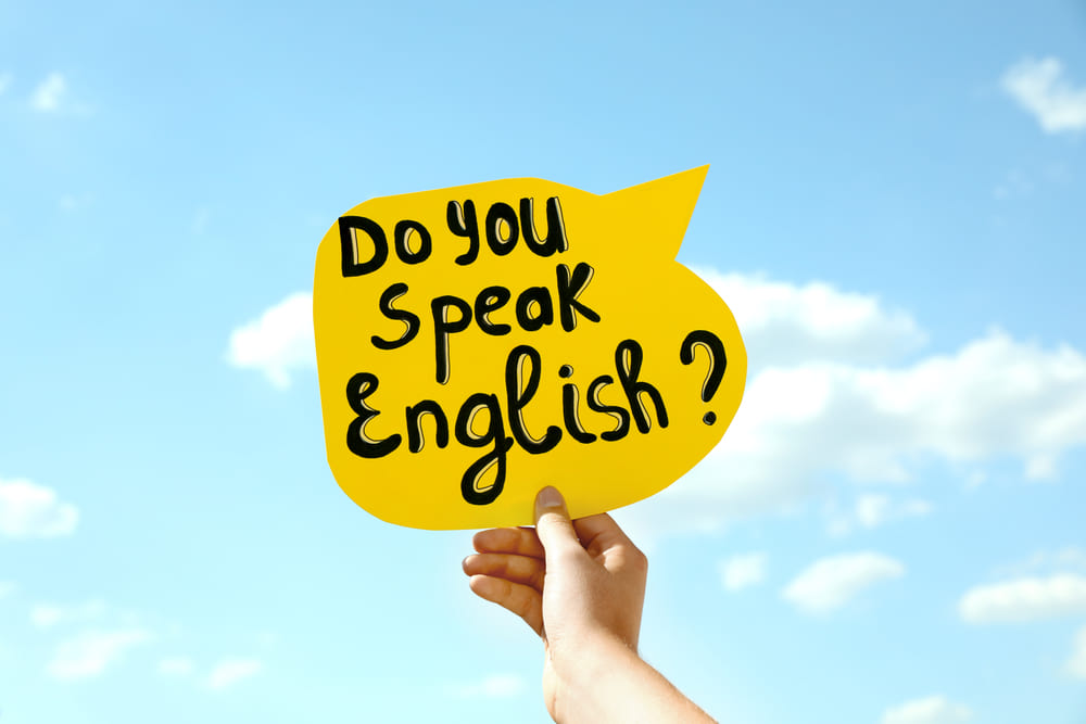 Cartaz amarelo com a frase “Do you speak English?” sendo segurado por uma mão contra o céu azul.