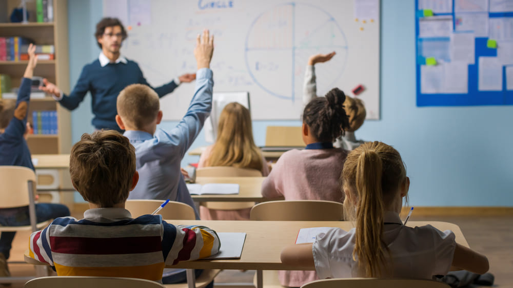 Crianças levantando as mãos em sala de aula, interagindo com o professor que está explicando no quadro.