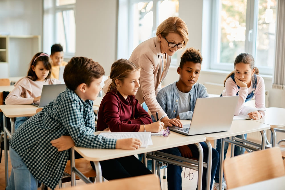 Professora orientando alunos no uso do computador em sala de aula, prática de letramento digital.