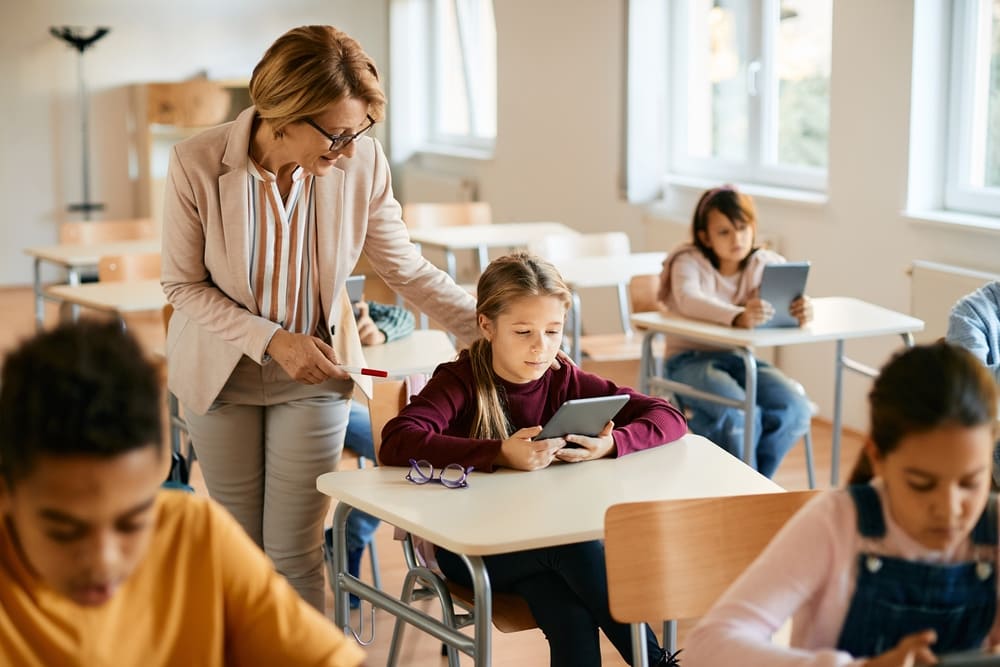 Professora ajudando aluna com tablet em sala de aula, exemplificando o uso da inteligência artificial na escola.