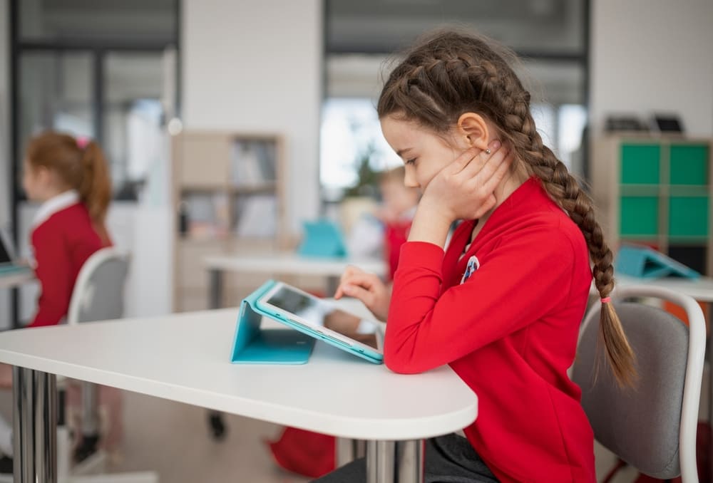 Aluna concentrada em tablet, com outros alunos ao fundo, representando o uso de inteligência artificial na escola no aprendizado.