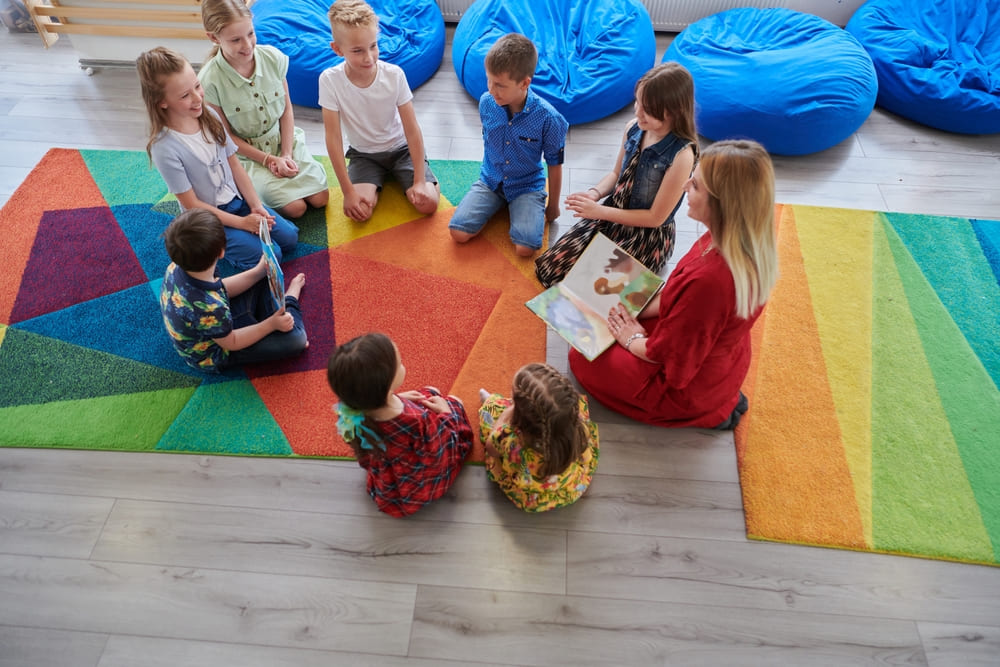 Professora lendo um livro para crianças em uma sala de aula infantil, com crianças sentadas ao redor em um tapete colorido.