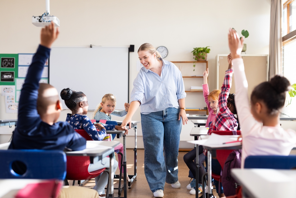 Professora interagindo com crianças em sala de aula, enquanto vários alunos levantam a mão para participar.