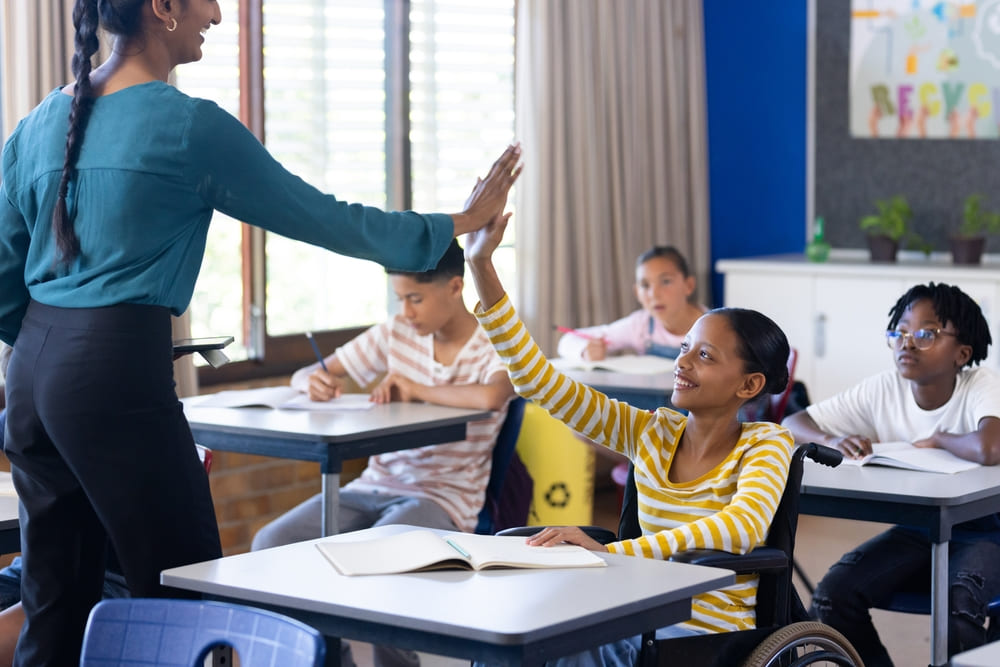 Criança em cadeira de rodas recebendo um high five da professora, exemplificando a inclusão através de tecnologia assistiva na educação.
