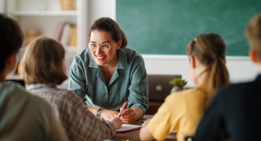 Professora na escola interagindo com seus alunos em sala de aula, com uma lousa verde ao fundo.