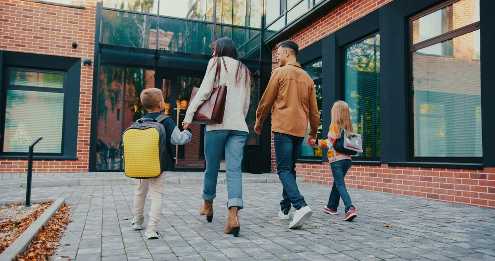 Pais de alunos caminhando com os filhos em direção à escola, simbolizando envolvimento familiar na educação.