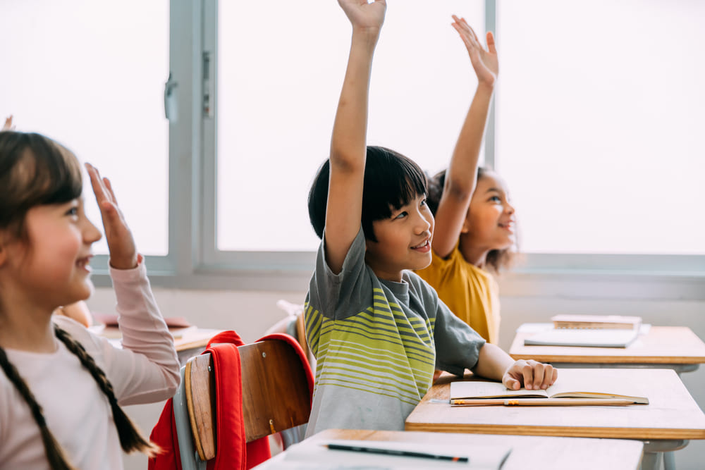Crianças levantando a mão para responder em sala de aula, um exemplo de participação ativa.