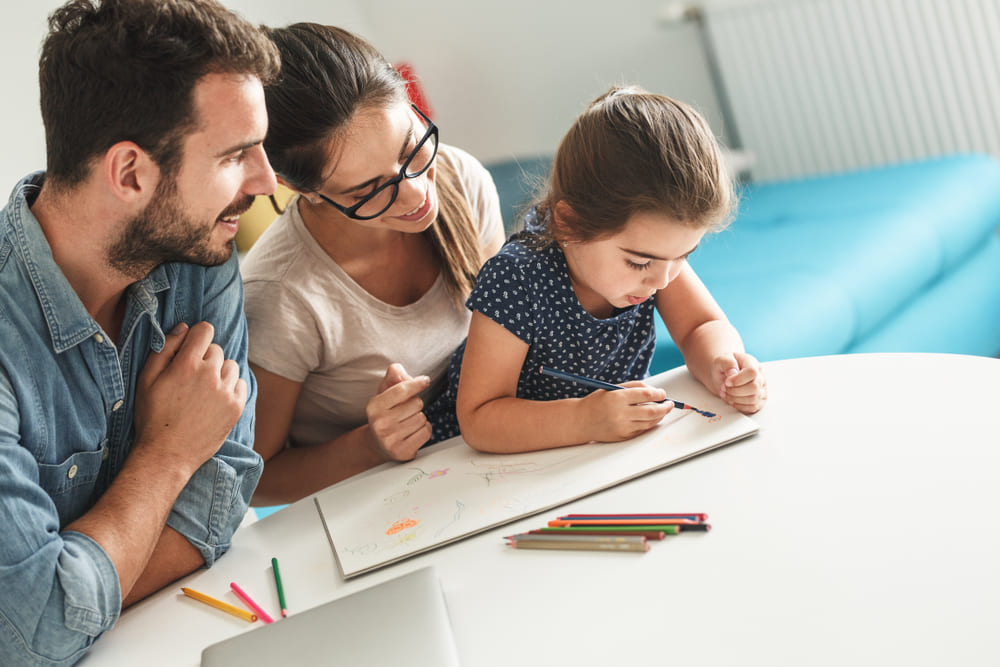 Pais auxiliando uma criança em uma atividade de desenho durante o recesso escolar, com lápis de cor sobre a mesa.